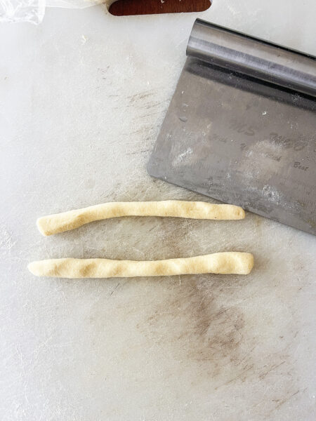 Ropes of semolina dough next to a bench scraper on a cutting board.