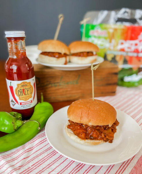 Sloppy joes made with Hatch chiles on a plate with peppers and ketchup behind it.