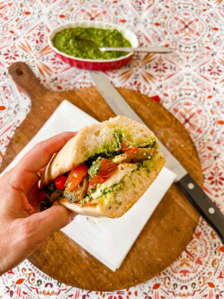 Hand holding tomato caprese sandwich over cutting board with pesto bowl.