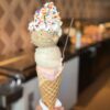 ‘Upcycled’ Ice Cream Gets New Flavors for 2025 Hand holding triple scoop ice cream cone with whipped cream and sprinkles.